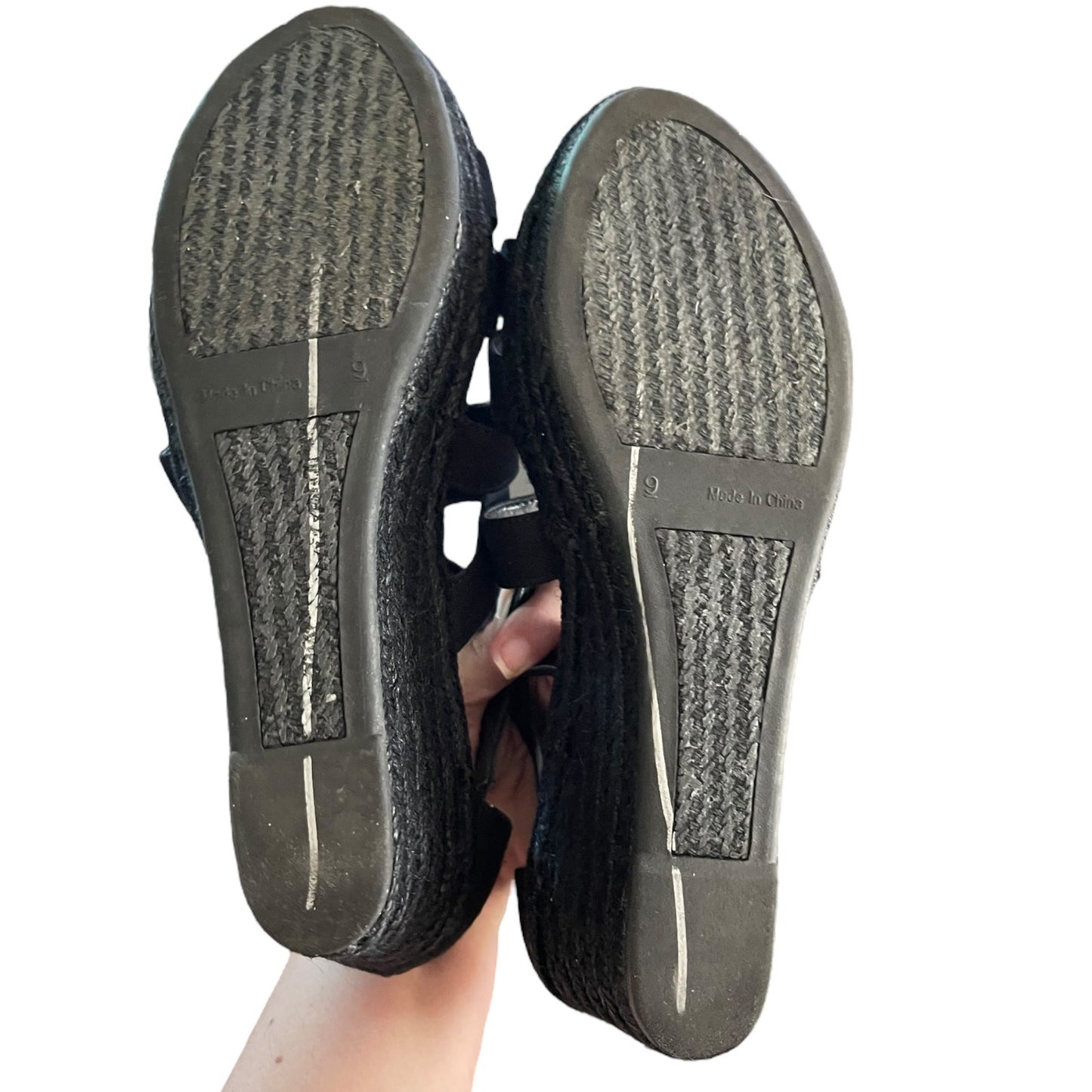 A pair of black espadrille sandals shown from the bottom, held up by a hand against a plain white background.