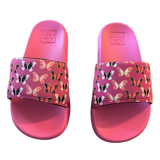 A pair of pink slide sandals with a wide strap covered in a repeating butterfly print. The brand name 'REEF' is embossed on the footbed of each sandal.