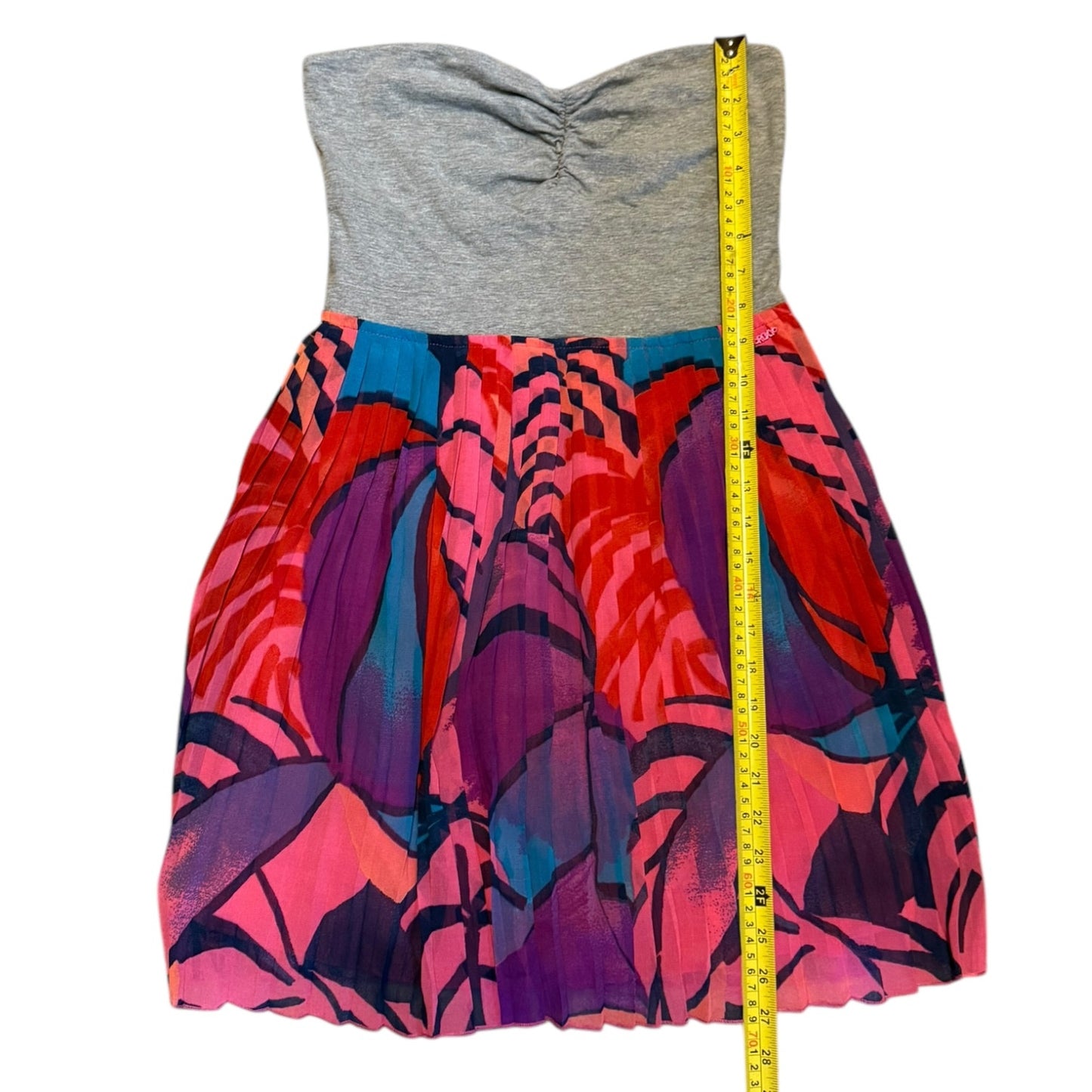 A strapless sweetheart dress with a heather gray ruched bodice and a vibrant, multicolored pleated skirt featuring pink, purple, red, and blue patterns. A yellow measuring tape is positioned vertically on the right side of the skirt to indicate its size.