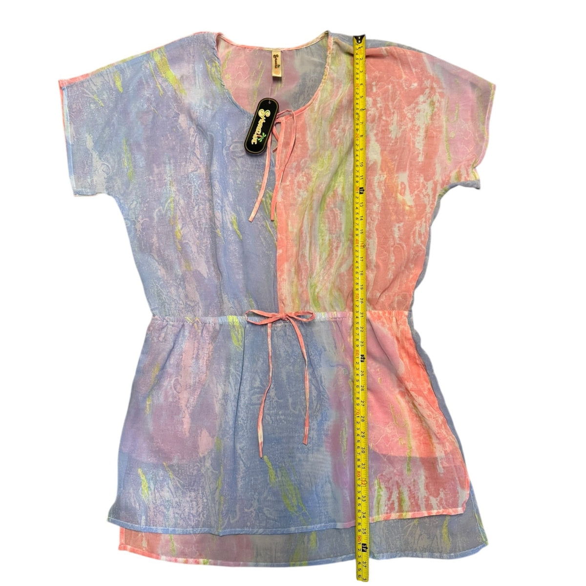 A short-sleeve tunic dress with a tie-dye pattern in pastel blue, pink, and orange, featuring a drawstring waist and a yellow measuring tape standing vertically next to it on a white background.