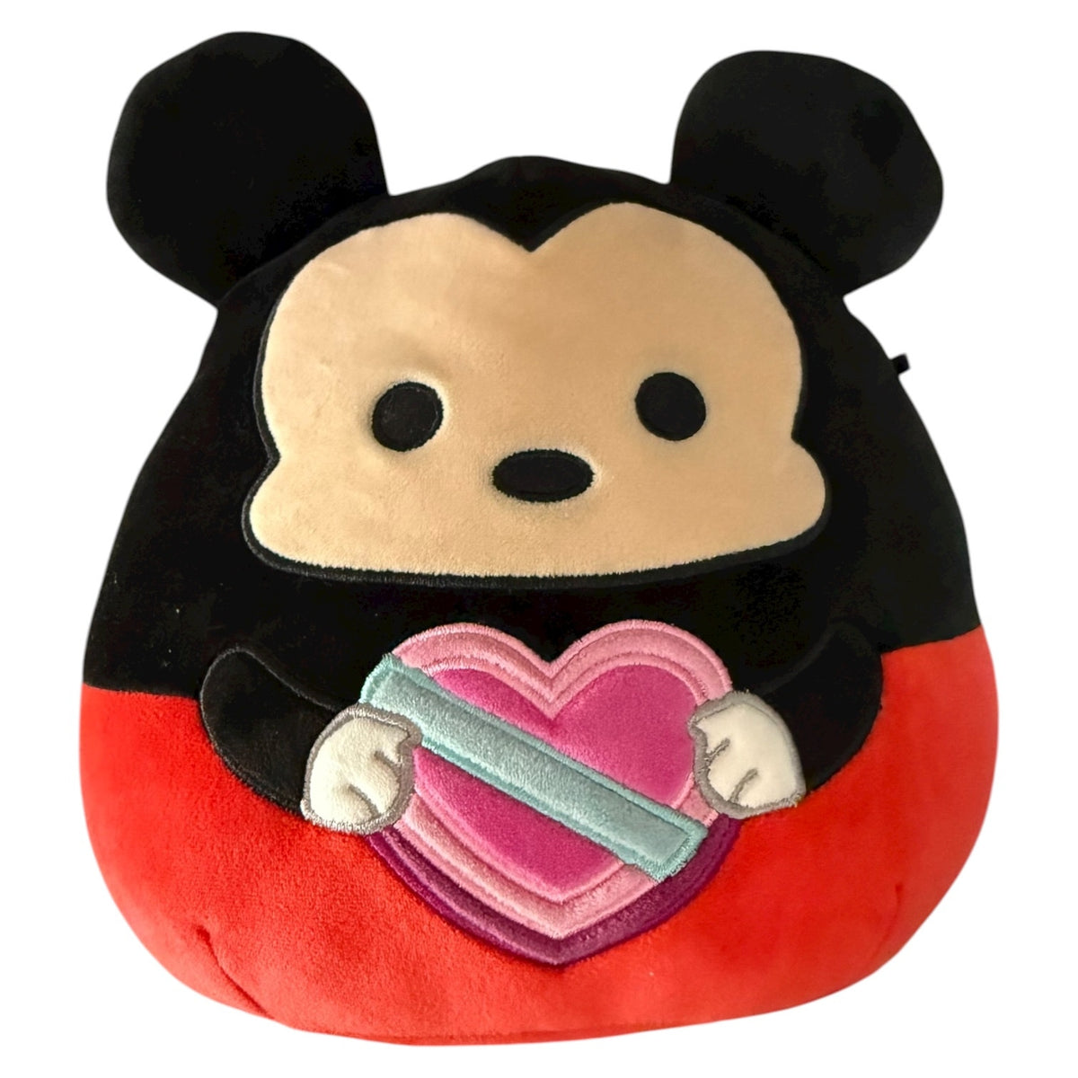 A plush Mickey Mouse plush with a black top section, tan face, red lower body, and a pink heart with a blue ribbon being held, shown on a plain white background.
