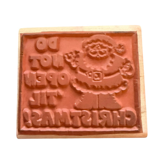 A wooden Christmas-themed rubber stamp featuring a raised Santa Claus figure and the text 'DO NOT OPEN TIL CHRISTMAS' in relief, shown on a plain white background.