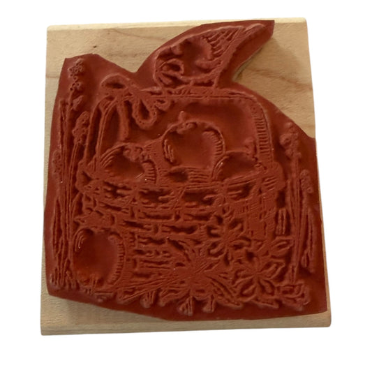 A wooden rubber stamp featuring an embossed autumn harvest design with apple, a basket, and a bird, shown on a light wood block against a white background.
