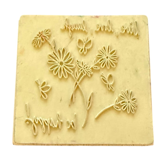 A square rubber stamp featuring embossed daisy flowers, leaves, butterflies, and cursive text, shown against a plain white background.
