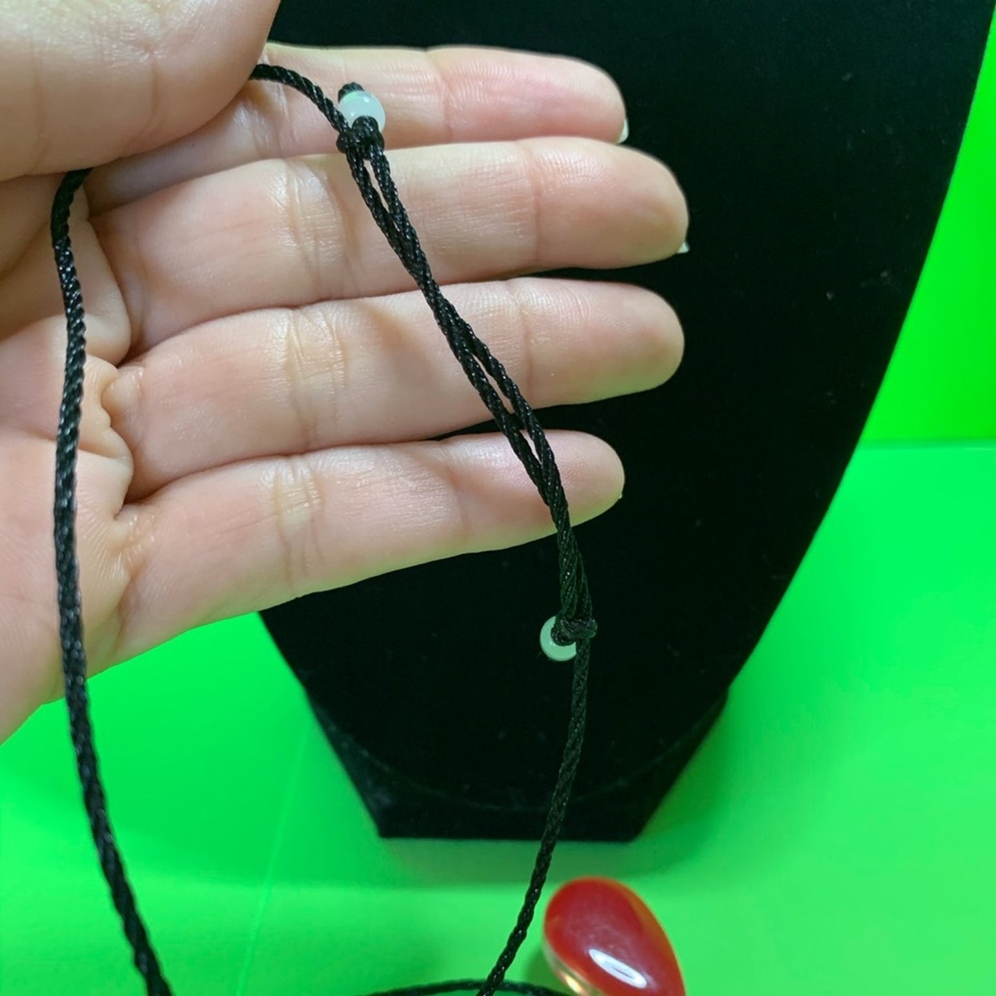 A close-up of a hand holding a black adjustable cord necklace with small white beads, positioned in front of a black display bust against a bright green background.