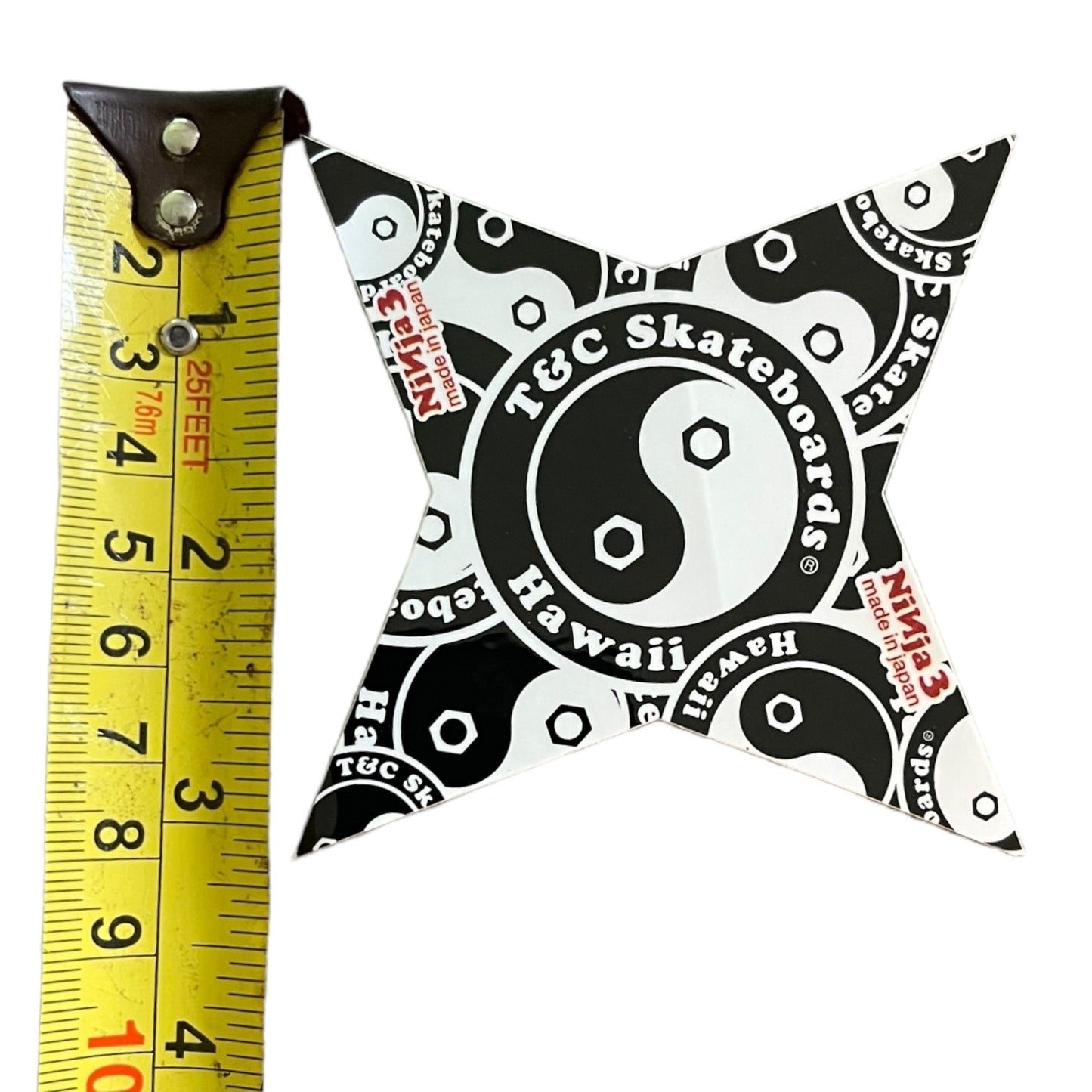 A black and white ninja star with a yin-yang and skateboarder pattern, shown next to a yellow tape measure to indicate its size. The pin cushion features the text 'T&C Skateboards Hawaii' and 'Skateboards Hawaii'.
