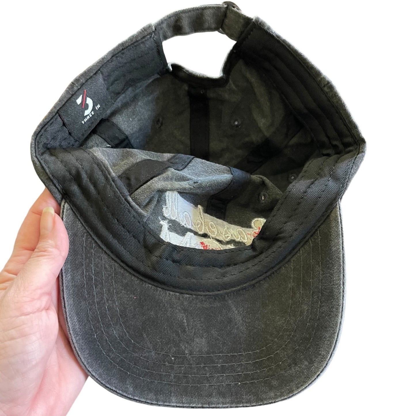 A dark gray black washed cotton baseball cap held open by a hand, showing the interior lining and adjustable strap.