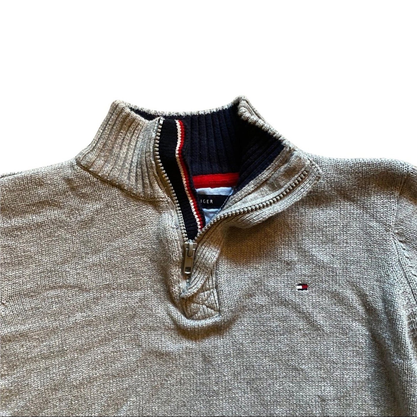 A close-up of a Tommy Hilfiger quarter-zip knit sweater in a gray color, showing the ribbed collar, zipper, and the signature red, white, and blue flag logo on the chest.