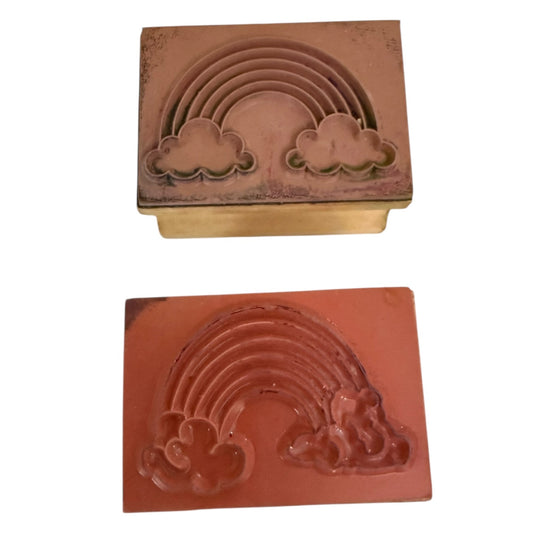 Two terracotta-colored rectangular rubber stamps with embossed rainbow and cloud designs, shown from opposite sides on a plain white background.