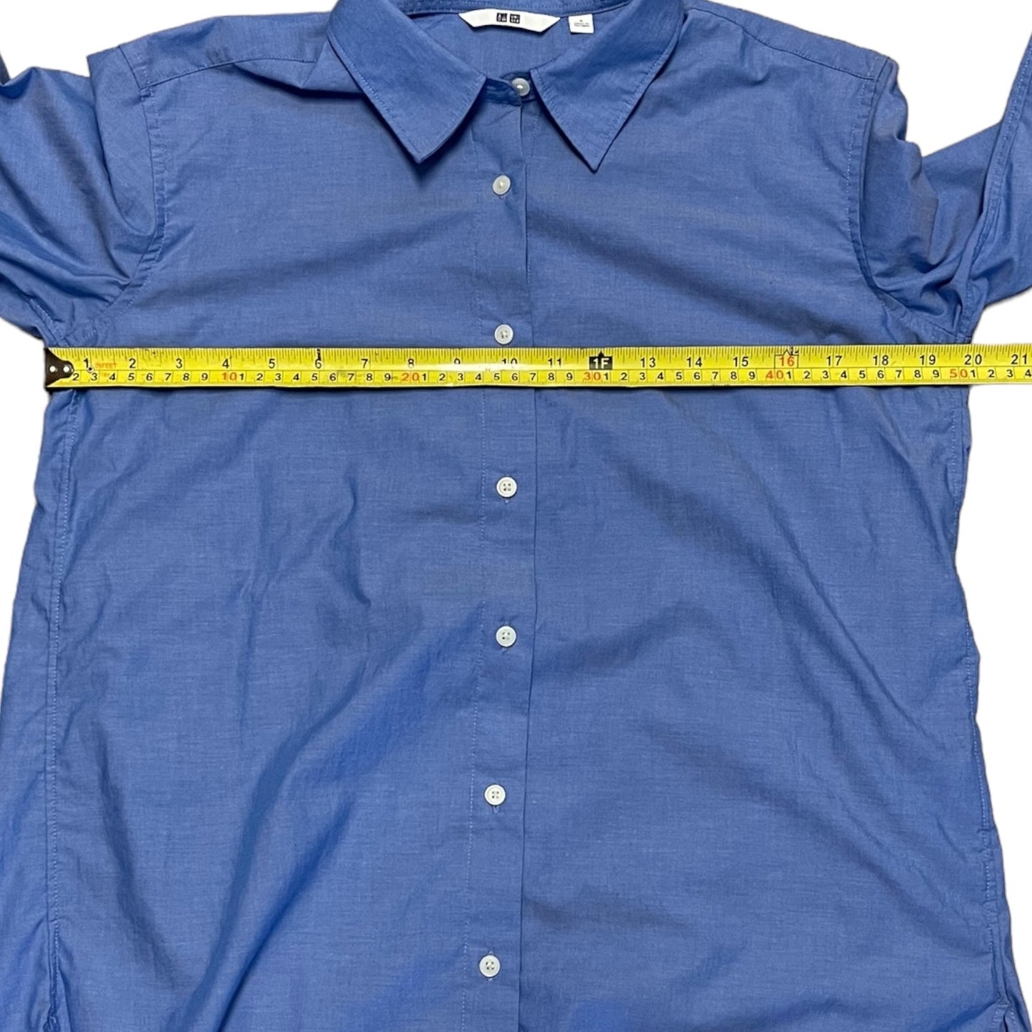 A blue long-sleeve button-up shirt laid flat on a white background, with a yellow tape measure stretched horizontally across the chest to show the width.