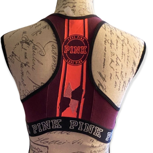 A sports bra shown from the back on a beige mannequin torso. The garment is burgundy with a vertical orange-red stripe down the center, a circular 'Love Pink' logo patch, and a black underband printed with 'PINK' in white letters.