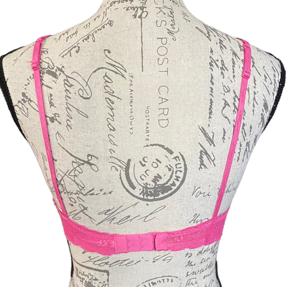 A bright pink lace bra displayed on a beige fabric dress form with a vintage postcard print. The back view shows thin pink shoulder straps and a ruffled pink underband.