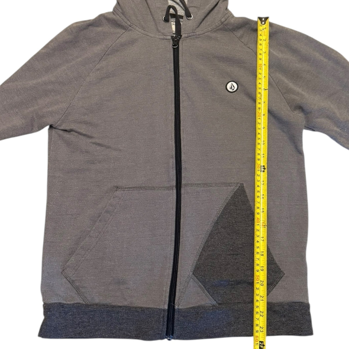 A gray zip-up hoodie with a black zipper, black drawstrings, and a small circular logo on the left chest, shown flat on a white background with a yellow tape measure standing vertically to indicate size.