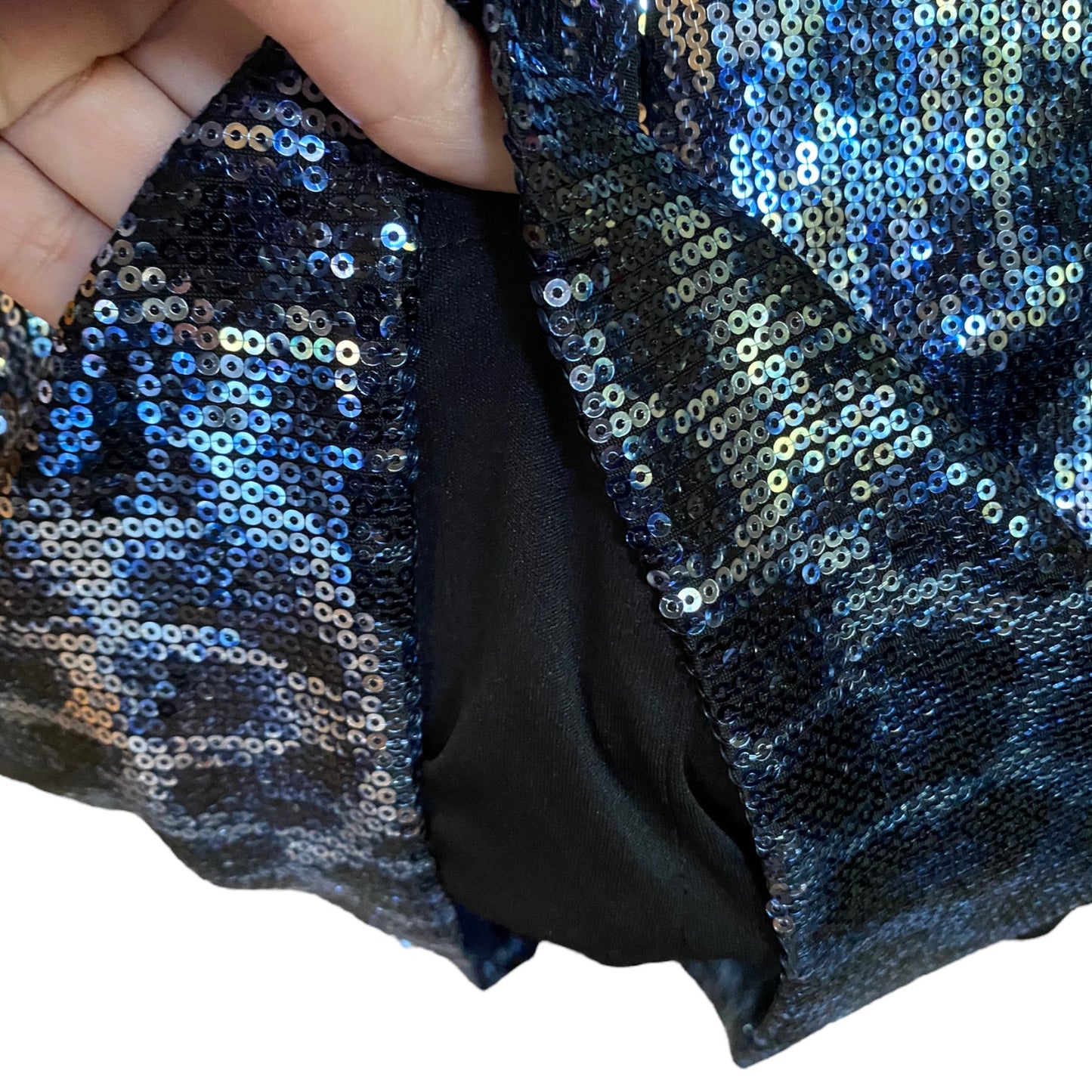 A close-up of a sequined fabric dress being held open by a hand, showing iridescent blue, silver, and black sequins over a black satin lining.