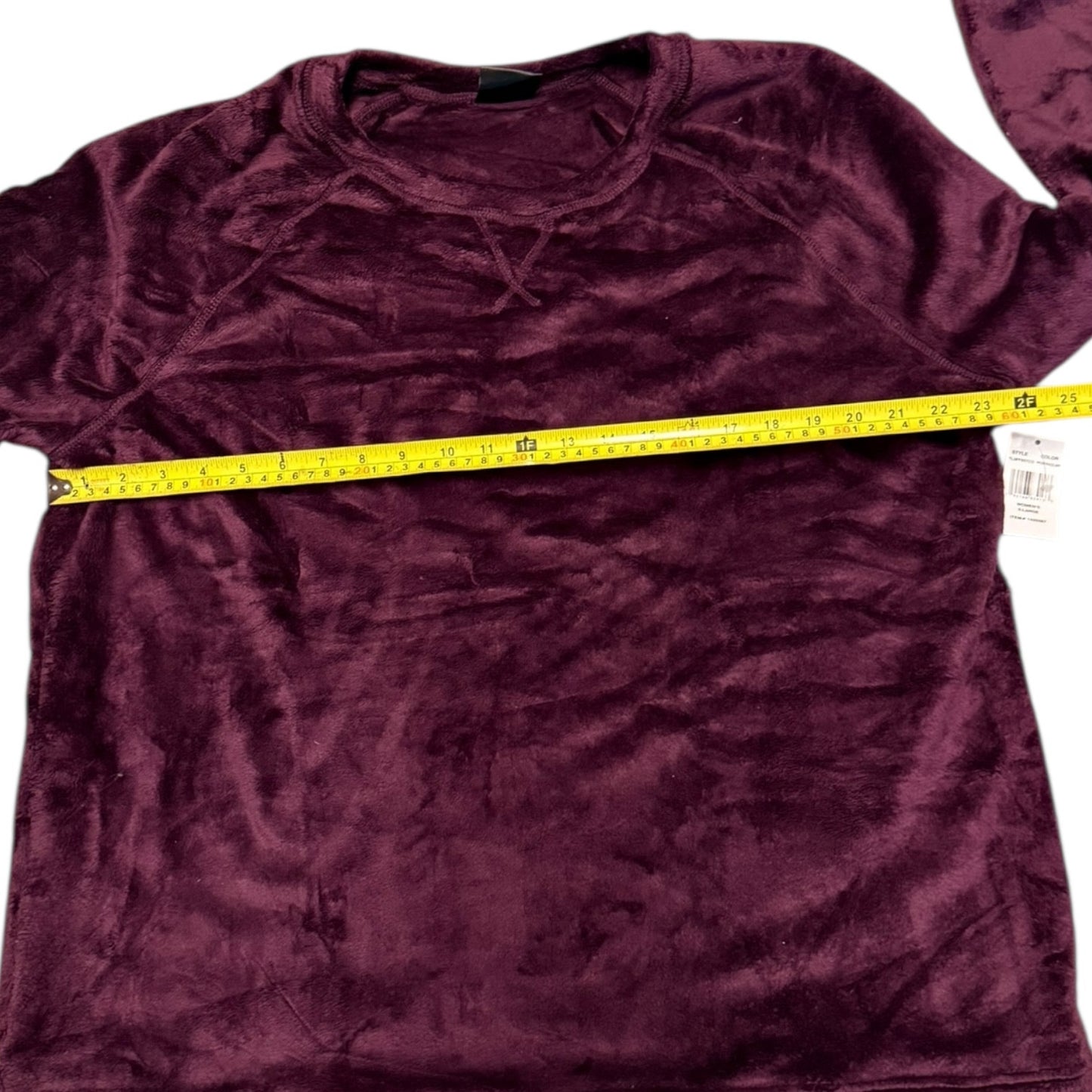 A plum purple plush fleece shirt laid flat on a white background, with a yellow tape measure stretched horizontally across the chest to show the width and a size tag visible on the right sleeve.