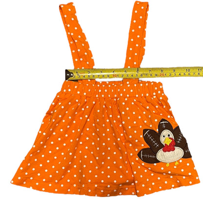 An orange and white polka dot baby/toddler skirt with straps, featuring a turkey applique on the lower right side and a yellow tape measure laid horizontally across the waistband to indicate size.