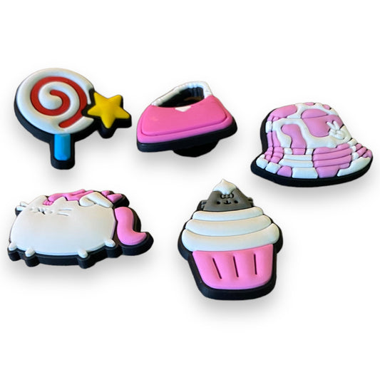 Five cute, 3D cartoon-style shoe charms with a llollipop with a star, a pink slipper, a pink-and-white bucket hat, a cupcake, and a white cat with a pink bow, all on a plain white background.