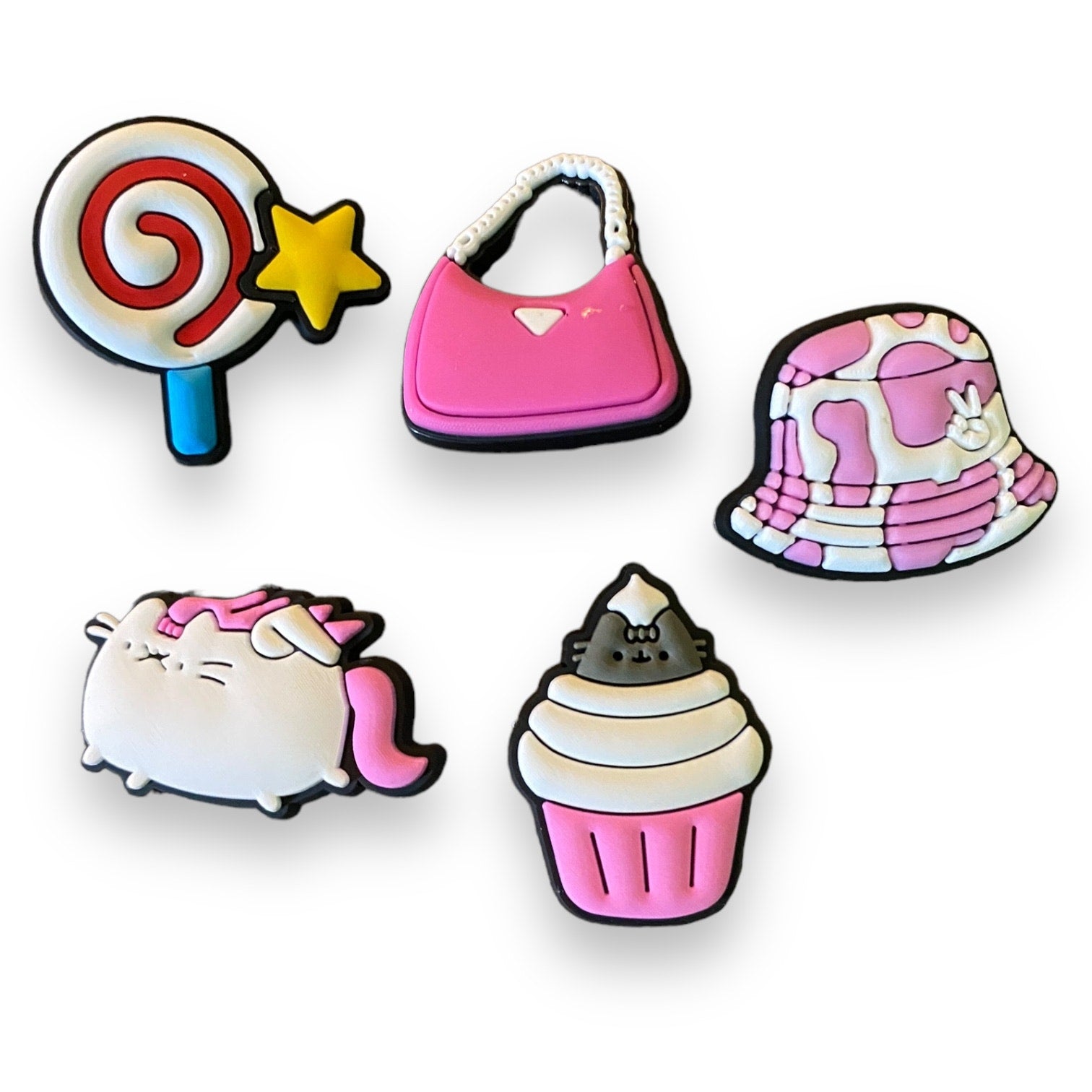 Set of colorful shoe charms including a lollipop, pink bag, cow print hat, cat with pink hair, and cupcake on a white background.