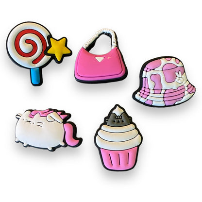 Set of colorful shoe charms including a lollipop, pink bag, cow print hat, cat with pink hair, and cupcake on a white background.
