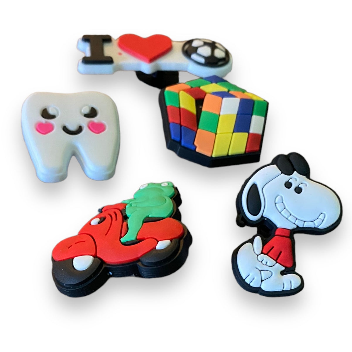A set of five colorful 3D cartoon-shaped croc charms on a white background, featuring Snoopy, a tooth with pink hearts, a Rubik's cube, a red scooter with a green frog, and a 'I <heart> soccer' design.