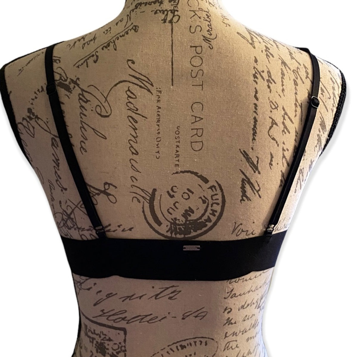 Black  bra shown from the back on a beige fabric dress form with a vintage postcard script print, against a white background.