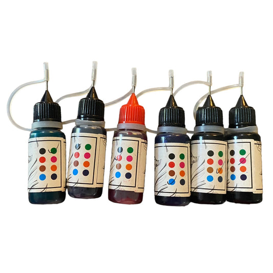 Six small tattoo ink bottles lined up on a white background, each with a black cap and a looped clear tube leading to a fine needle applicator. 