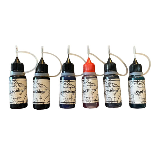 Six small tattoo ink bottles lined up on a white background, each with a black cap and a looped clear tube leading to a fine needle applicator. 