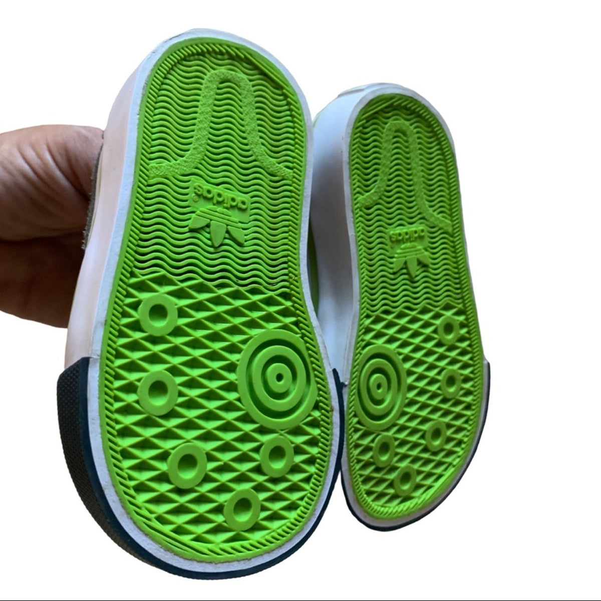 A close-up of a person's hand holding a pair of Adidas sneakers to show the textured green outsoles, with the Adidas trefoil logo embossed on the bottom of the soles.