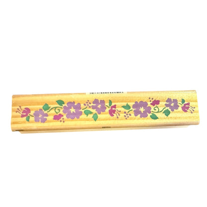 A rectangular wooden stamp with a light natural wood finish, decorated with a horizontal floral pattern of purple hibiscus flowers and green leaves, shown against a plain white background.