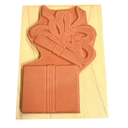 A terracotta-colored rubber stamp featuring a gift box with a ribbon and bow design, mounted on a light-colored wooden block against a white background.
