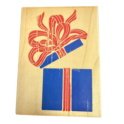 A wooden rubber stamp with open gift box with a red ribbon and bow above two blue gift boxes tied with red ribbons, set against a plain white background.