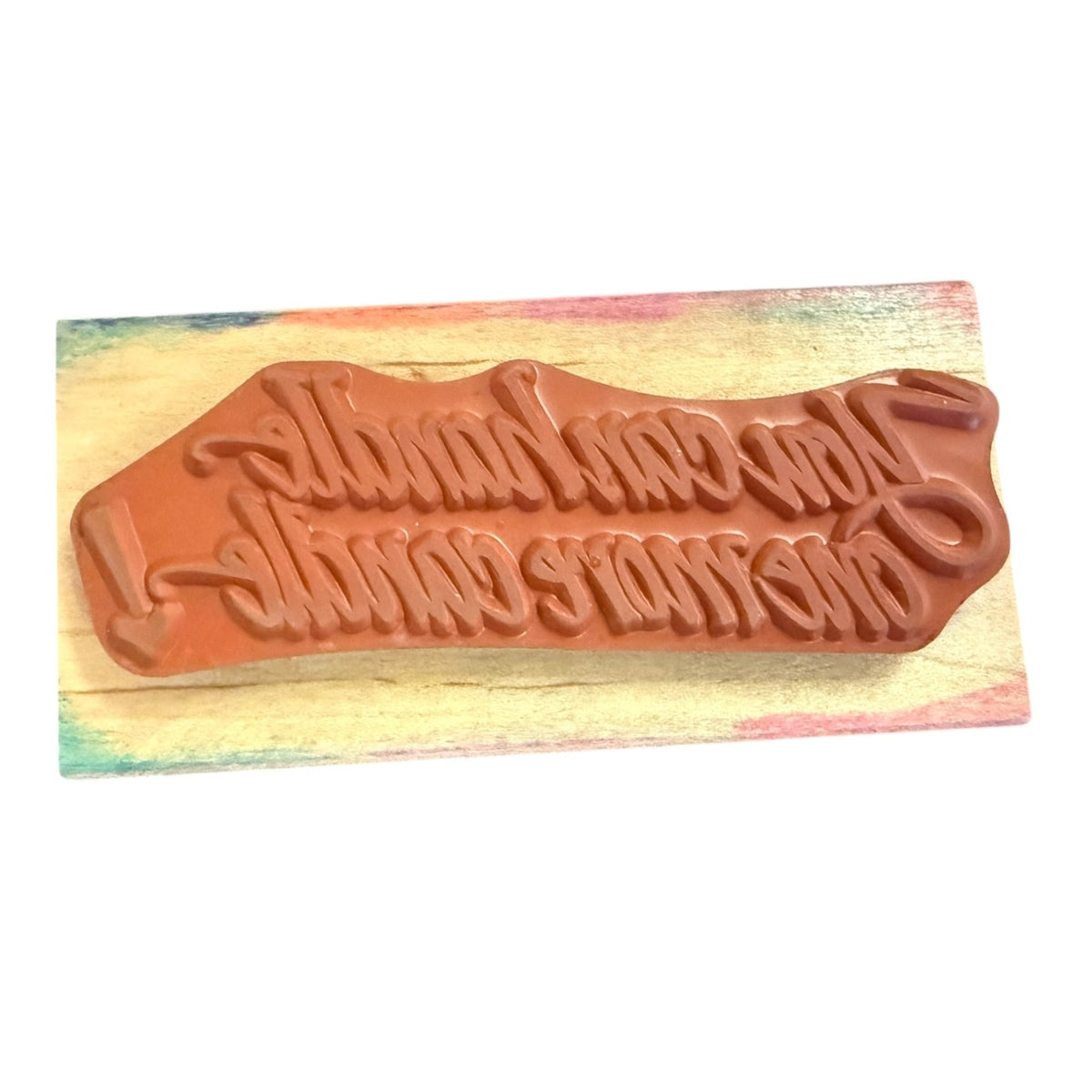 A terracotta-colored rubber stamp embossed in stylized script, displayed on a colorful painted wooden block against a white background.