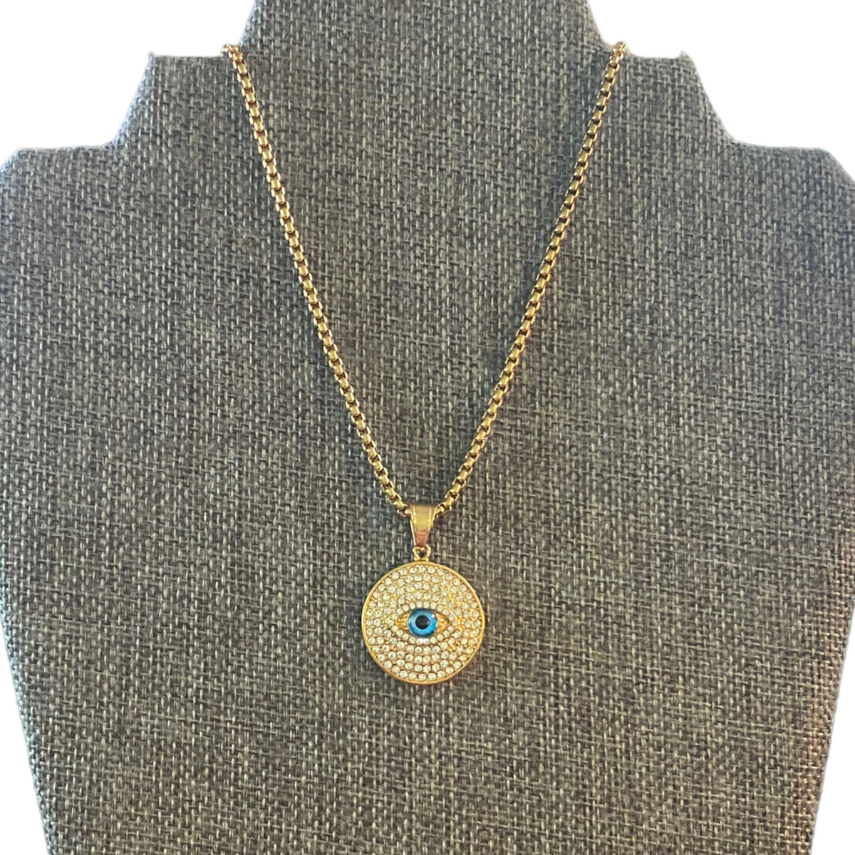 A gold-toned necklace with a circular evil eye pendant displayed on a gray fabric jewelry bust. The pendant features a textured gold background, a border of small clear stones, and a central blue eye detail.
