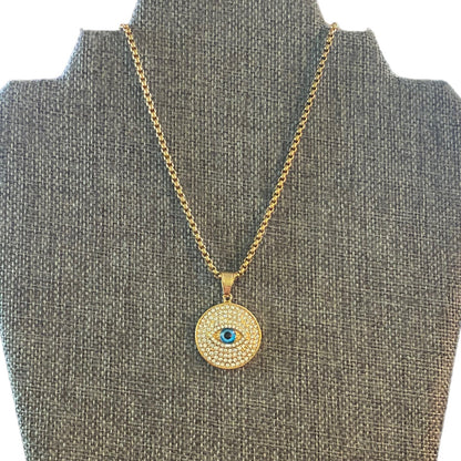 A gold-toned necklace with a circular evil eye pendant displayed on a gray fabric jewelry bust. The pendant features a textured gold background, a border of small clear stones, and a central blue eye detail.