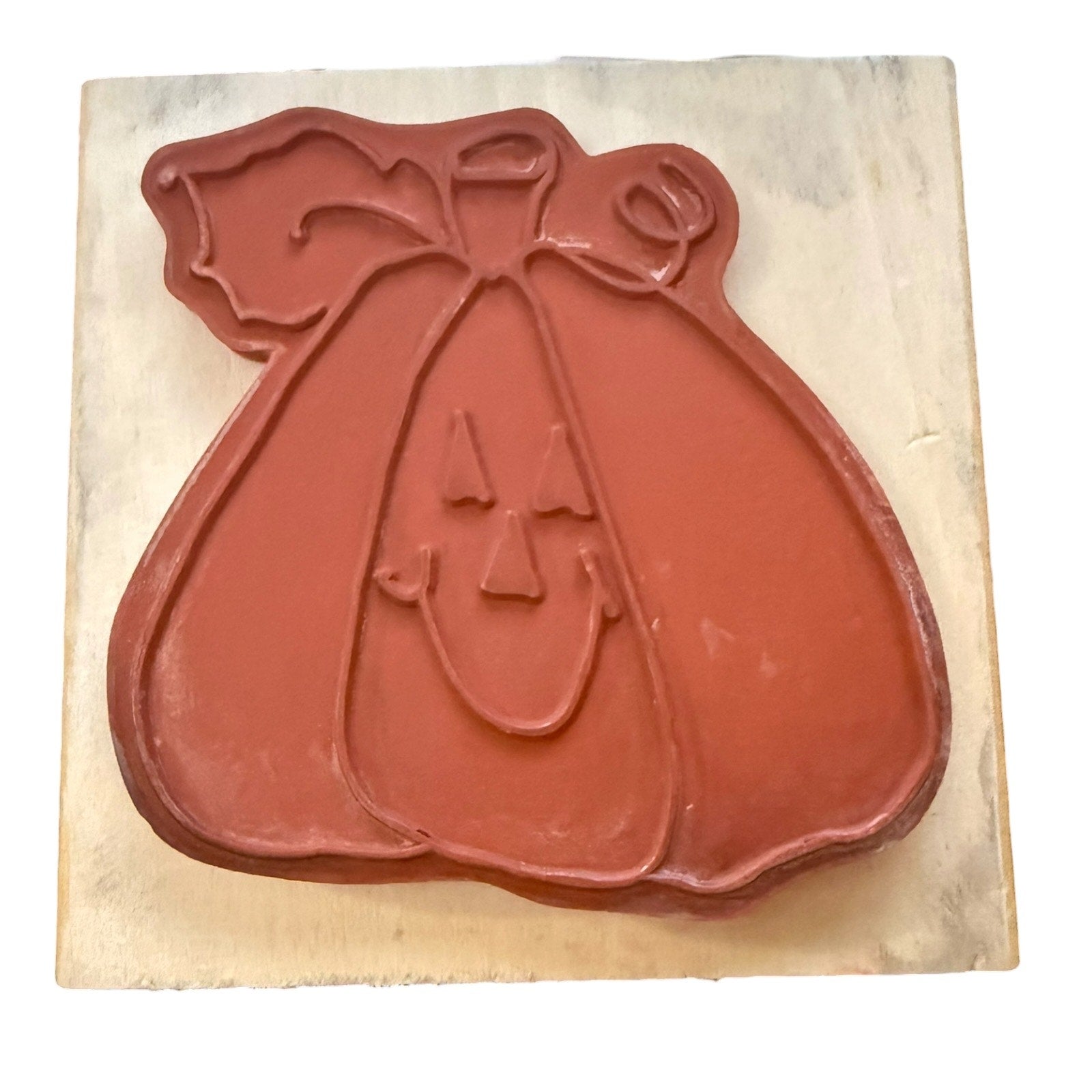 A pumpkin-shaped stamp or mold with a carved jack-o'-lantern face, mounted on a square light beige wooden block against a white background.