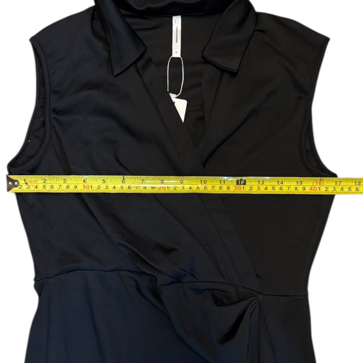 A black sleeveless collared wrap-style dress laid flat on a white background, with a yellow tape measure stretched horizontally across the chest to show the width.