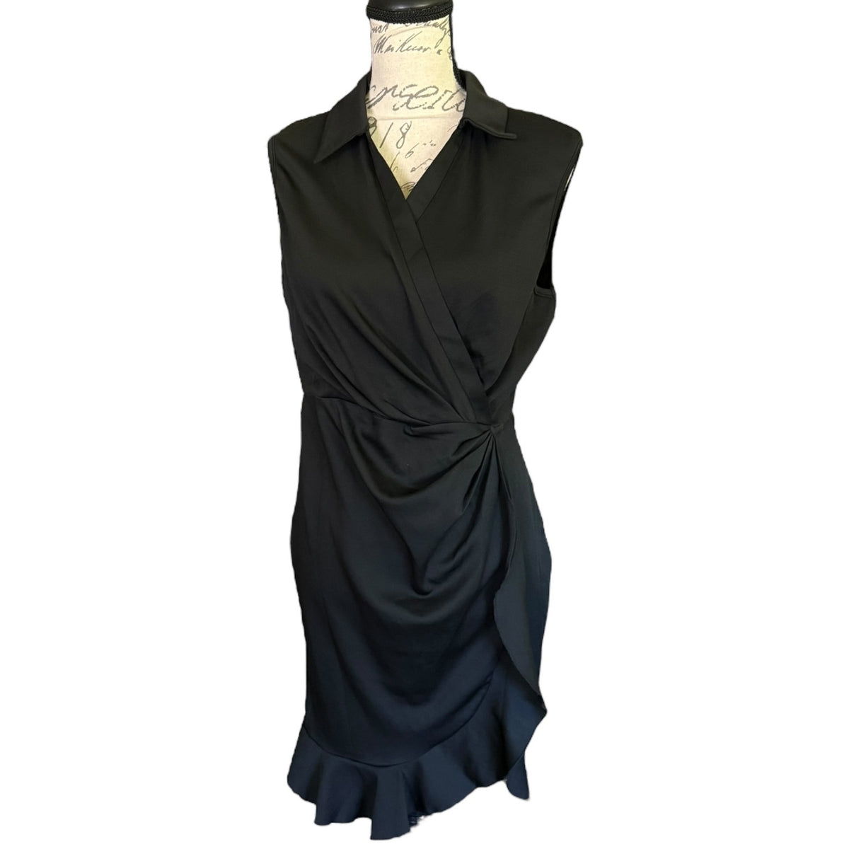 A sleeveless black faux wrap-style dress with a collar and gathered ruffle detail at the waist, displayed on a dress form against a plain white background.