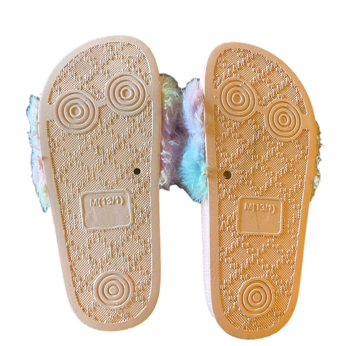 A pair of plush slippers shown from the sole side, featuring a textured pink rubber outsole with circular tread patterns and a fluffy pastel rainbow lining peeking out from the sides.