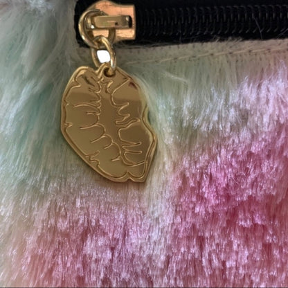 Close-up of a gold-toned zipper pull charm shaped like a stylized lips , attached to a black zipper on a fuzzy pastel surface with mint, pink, and lavender tones.