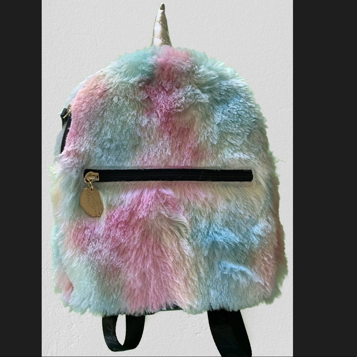 A fluffy faux-fur backpack with a pastel rainbow pattern of pink, blue, multicolor featuring a silver horn on top, a black horizontal zipper, and a gold charm.
