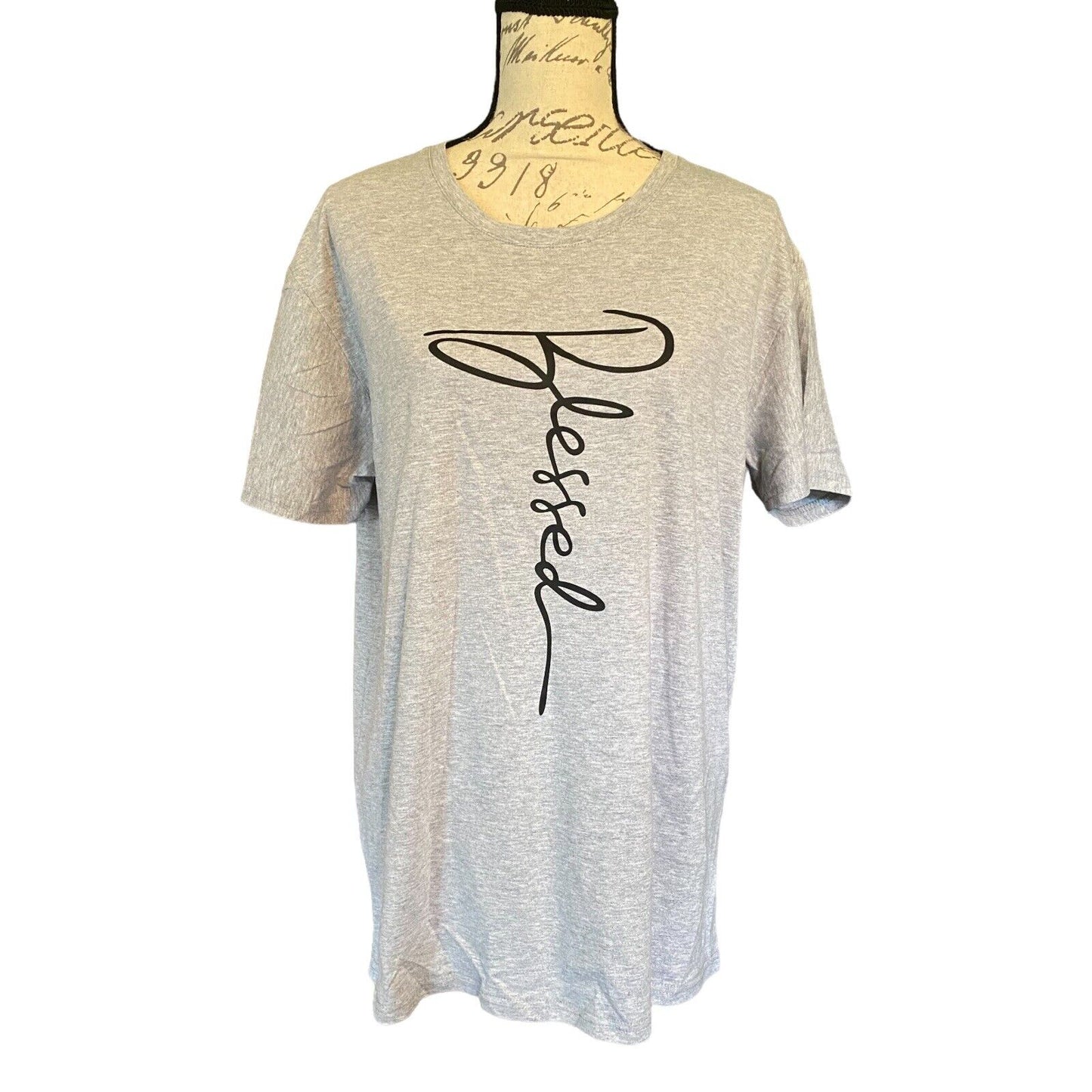 A light heather gray short-sleeve t-shirt . The shirt features a large black cursive script graphic spelling 'Blessed' vertically on the front.