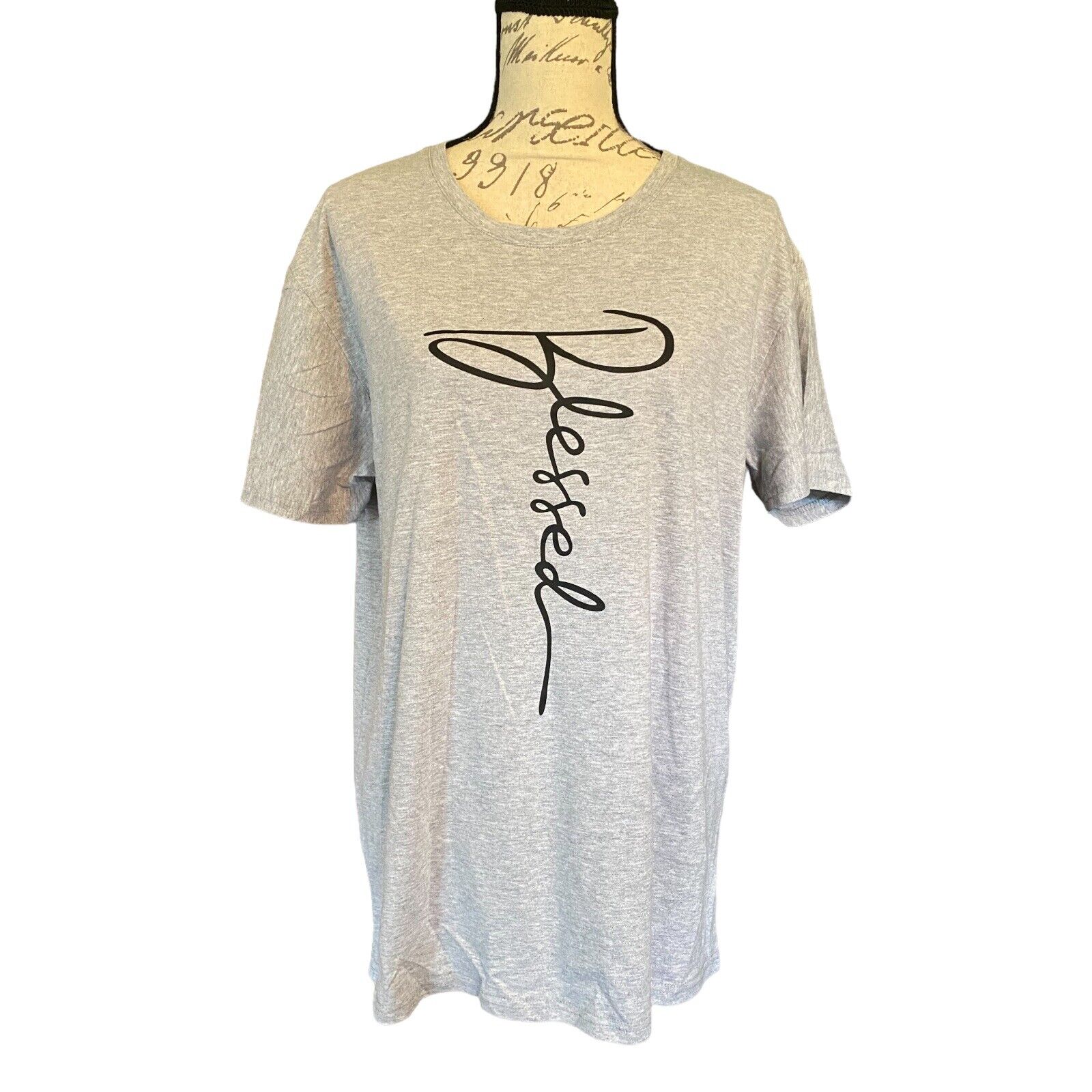 A light heather gray short-sleeve t-shirt . The shirt features a large black cursive script graphic spelling 'Blessed' vertically on the front.