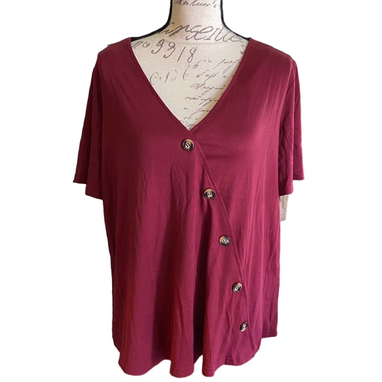 A women's short-sleeve top in maroon, displayed on a vintage-style dress form with cursive writing on the front. The top features a V-neckline and a front button closure with dark buttons.