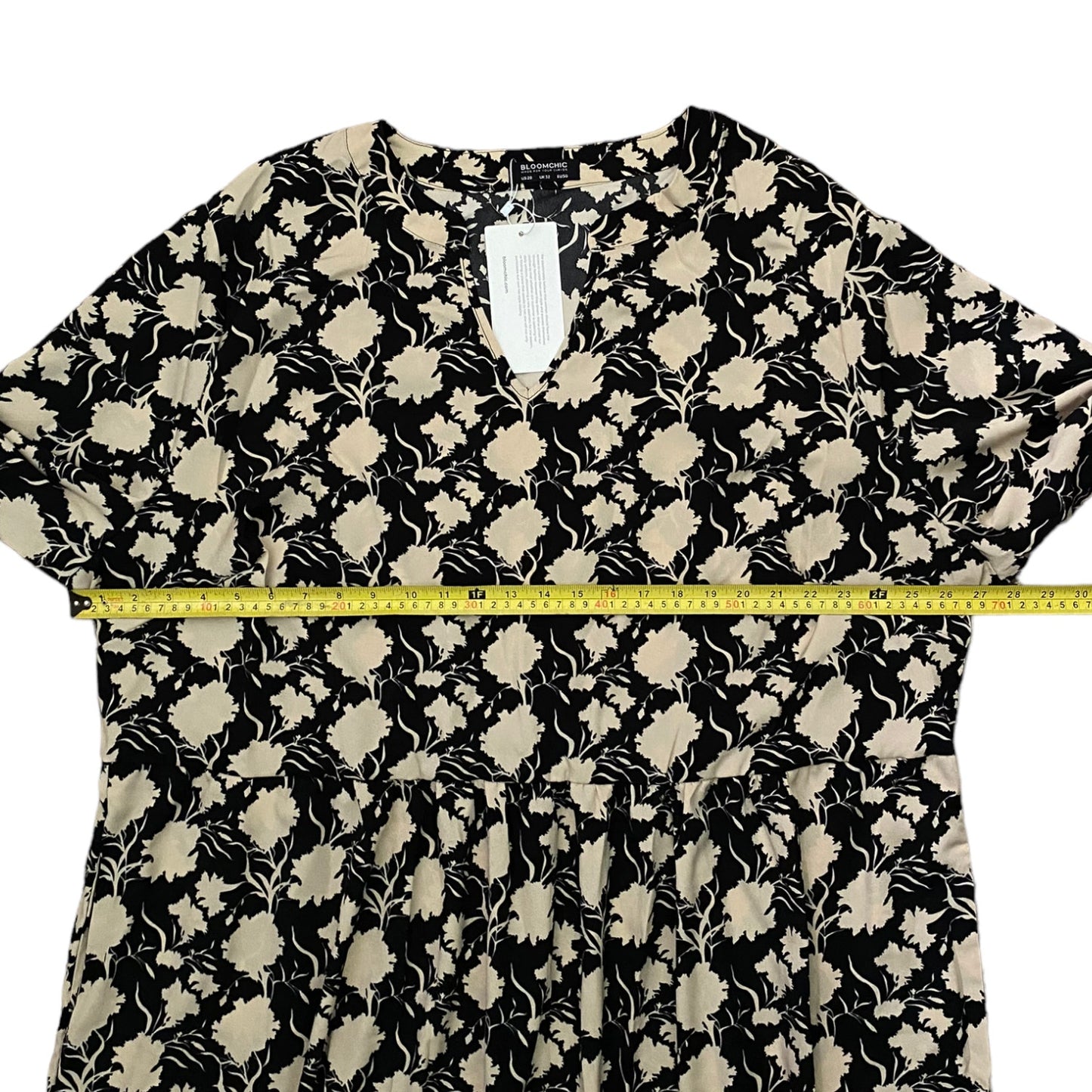 A black and beige floral print dress laid flat on a white background, with a yellow measuring tape stretched horizontally across the waist area to show the width.