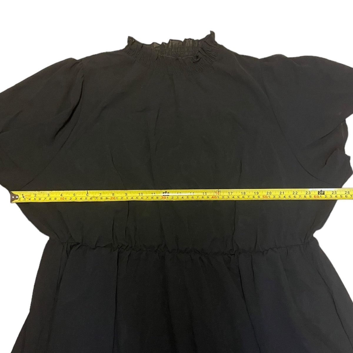 A black dress laid flat on a white background with a yellow tape measure stretched horizontally across the waist area to show the garment's measurements.