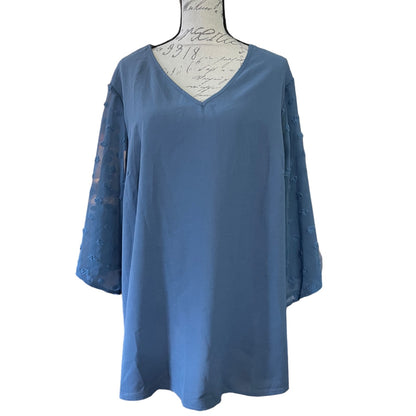 A dusty blue tunic top with three-quarter sleeves, featuring sheer lace sleeves with embroidered floral details and a V-neckline, displayed on a white dress form against a white background.