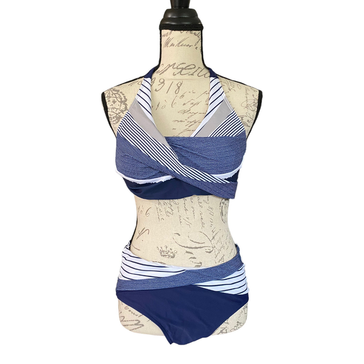 A two-piece bikini set displayed on a vintage-style beige mannequin torso. The set features a halter-style top with a criss-cross wrap design in navy blue, medium blue, and white striped fabric, paired with matching high-waisted bottoms in navy blue and white striped fabric.