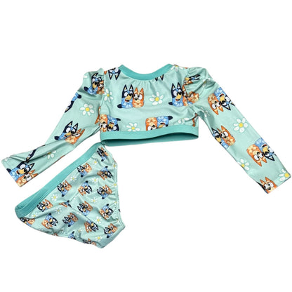 A two-piece children's swim set featuring a long-sleeve top and matching bottoms in a light mint green color, both printed with cartoon dogs and white daisies. 