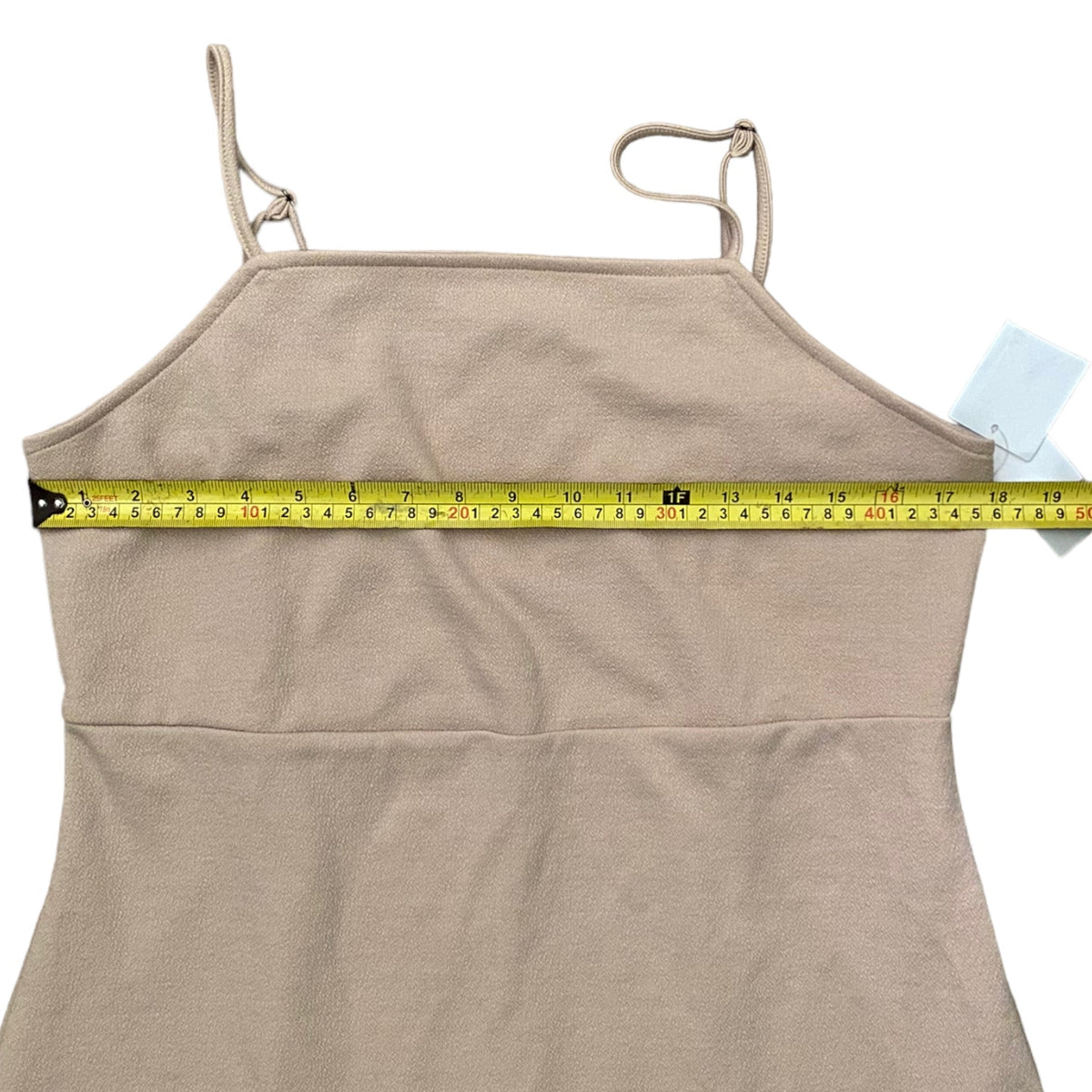 A beige spaghetti-strap dress with a yellow tape measure stretched horizontally across the chest to show the bust measurement, on a white background.