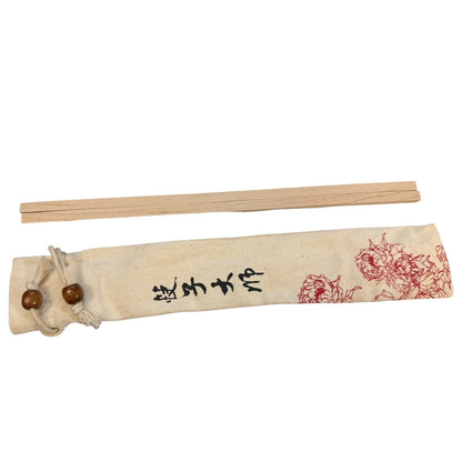 A pair of light wood chopsticks placed above their fabric storage sleeve. The sleeve is off-white with black characters and red floral illustrations, tied with a cord and two wooden beads.